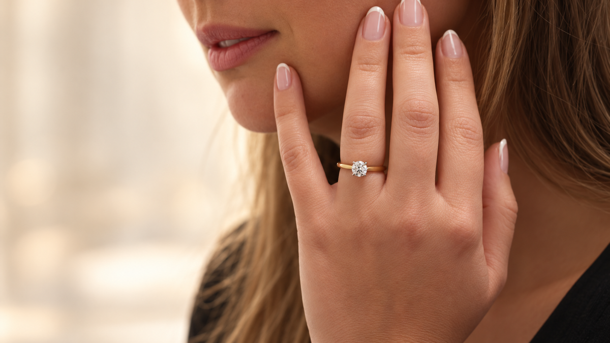 Yellow gold solitaire diamond ring worn on a hand with a French manicure, resting gently against the model's face.